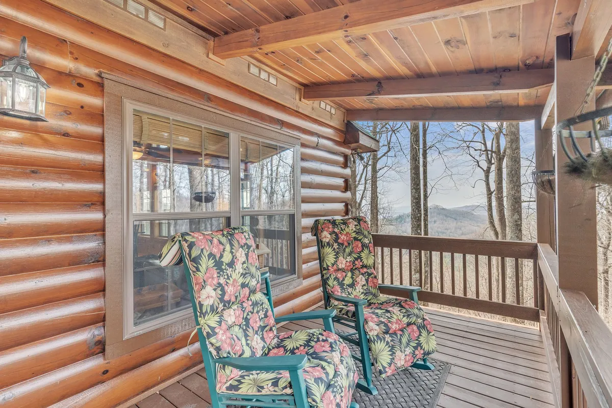 An inviting front porch with floral-patterned chairs is set against the rustic log cabin walls, providing a cozy space to enjoy the fresh mountain air. Through the windows, glimpses of the lush landscape enhance the sense of immersion in nature. The porch swing is a focal point for enjoying morning coffee with panoramic views.
