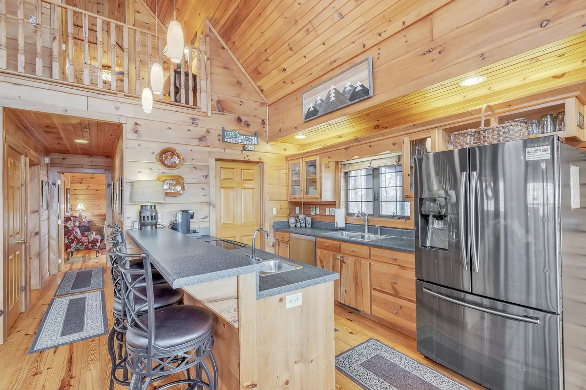 A bright, open kitchen featuring a spacious island with bar stools, stainless steel refrigerator, and wooden cabinetry. Large windows offer abundant natural light and scenic views, with the kitchen seamlessly connecting to living spaces.