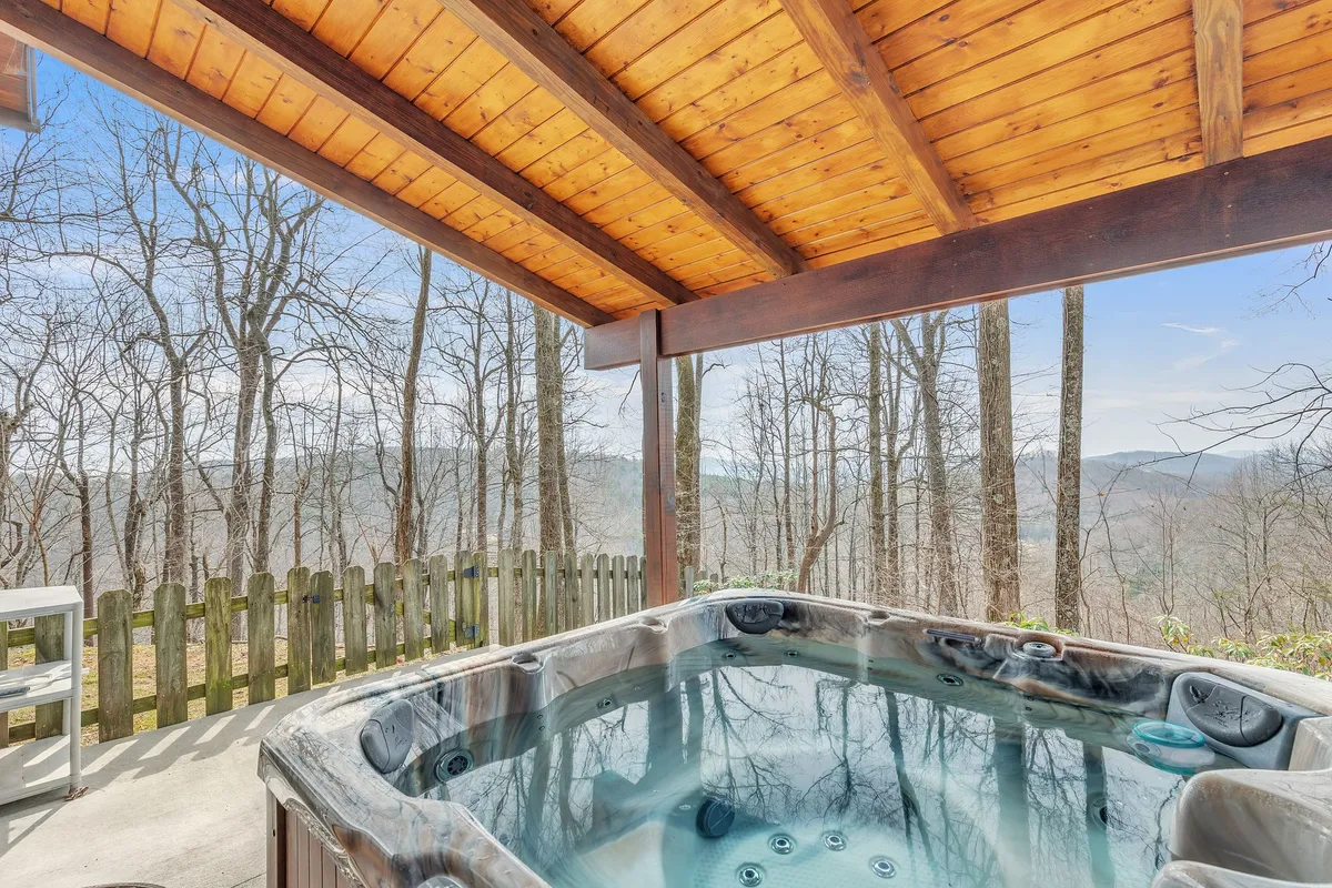 This exterior image features a private hot tub nestled under a wooden pavilion, surrounded by mature trees and offering a sweeping view of the Blue Ridge Mountains. The tranquil setting creates an atmosphere of seclusion and relaxation, ideal for couples seeking a romantic retreat amid nature.