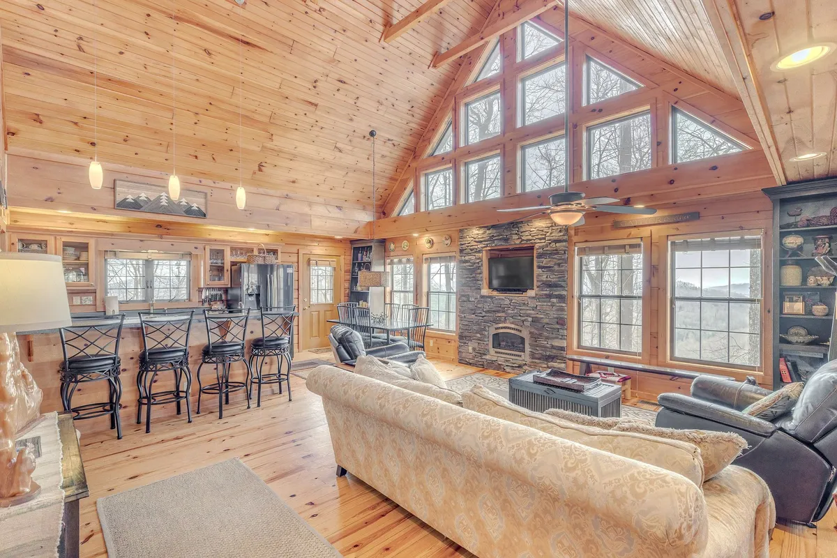 A spacious, inviting living area with large windows showcasing the surrounding landscape, a stone fireplace for cozy gatherings, a well-appointed bar area, and ample comfortable seating arranged for easy conversation or group activities.