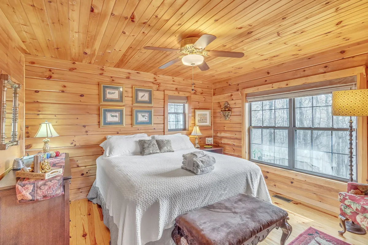 This image showcases a cozy bedroom with a bed dressed in white quilted linens, wood-paneled walls, a large window allowing abundant natural light, and a decorative lamp and artwork. The setting feels warm and restful, inviting relaxation and comfort.